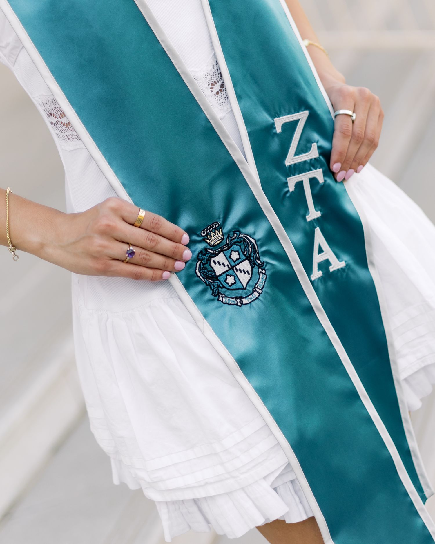 Close-up of a ZTA sorority graduation sash in teal with embroidered letters and crest on a white dress.