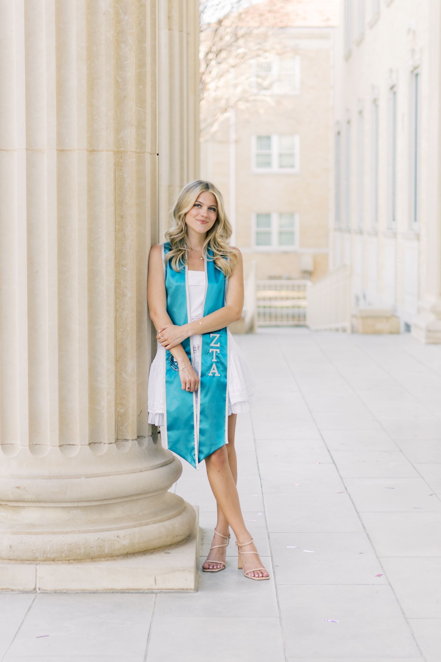 Blonde woman in white dress with teal ZTA graduation stole leaning against column at TCU.