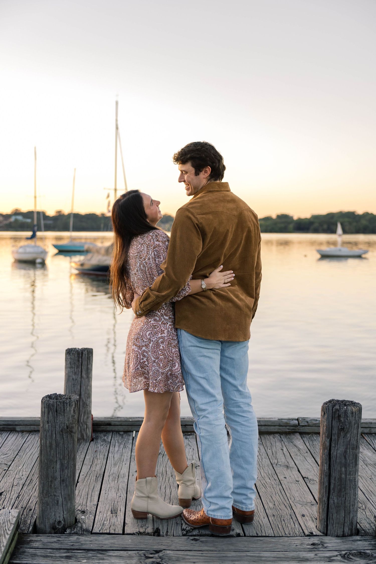 Couple embracing on a dock at sunset, gazing at each other with sailboats on the lake behind them.