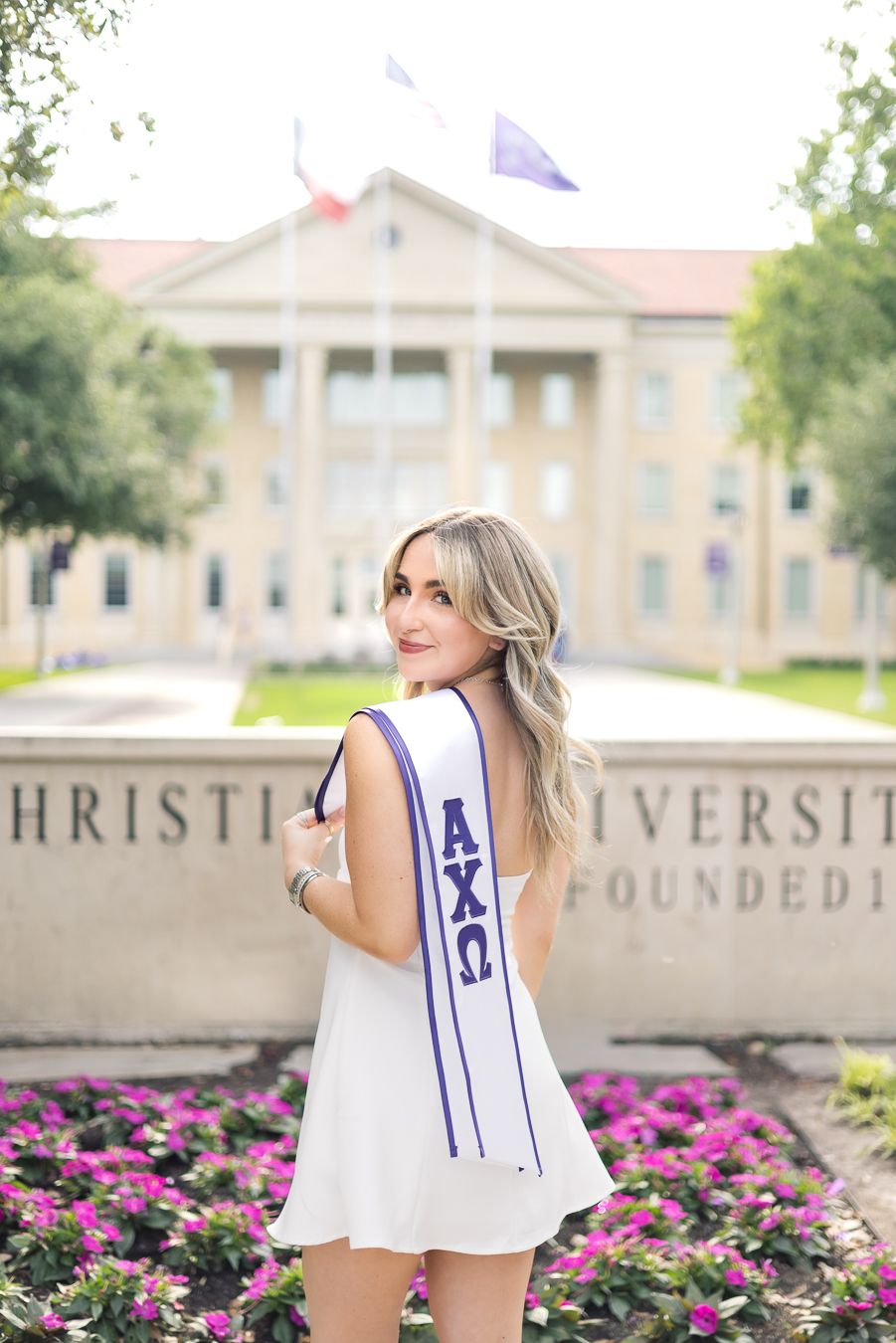 Young woman in white dress wearing Alpha Chi Omega sash poses in front of Texas Christian University sign.