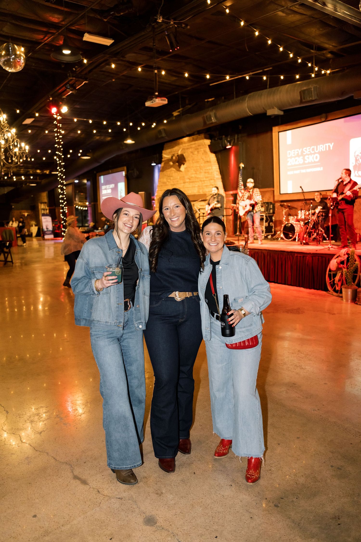 Three women smile at an indoor event venue with string lights, a live band on stage, and rustic decor.