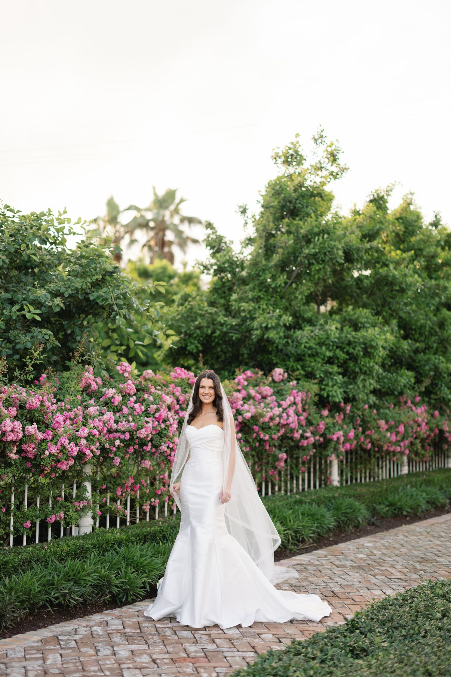 Bride in white mermaid gown and veil poses on brick path before pink roses and lush greenery.