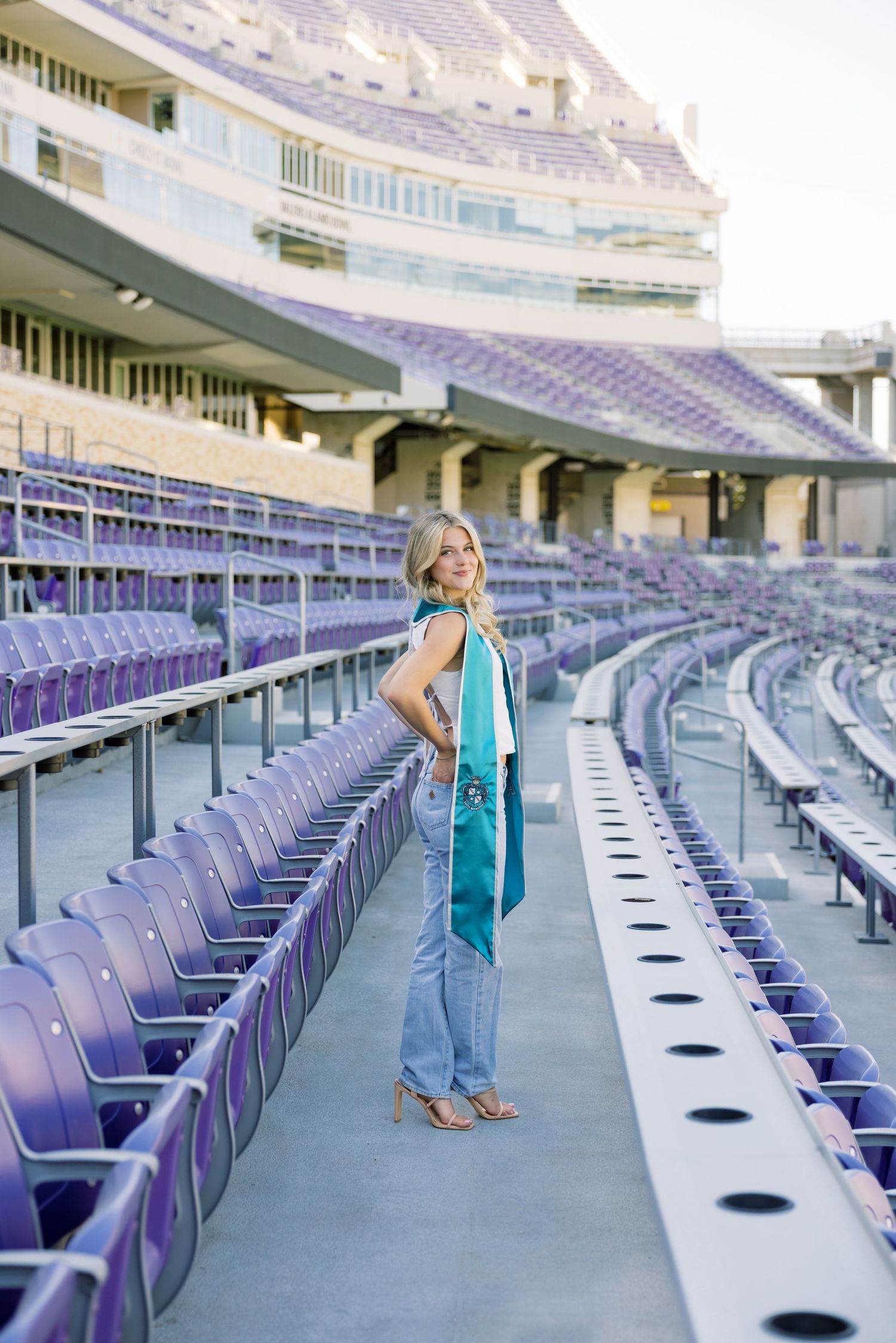 Blonde woman in jeans and white top with teal graduation stole standing in purple TCU stadium.
