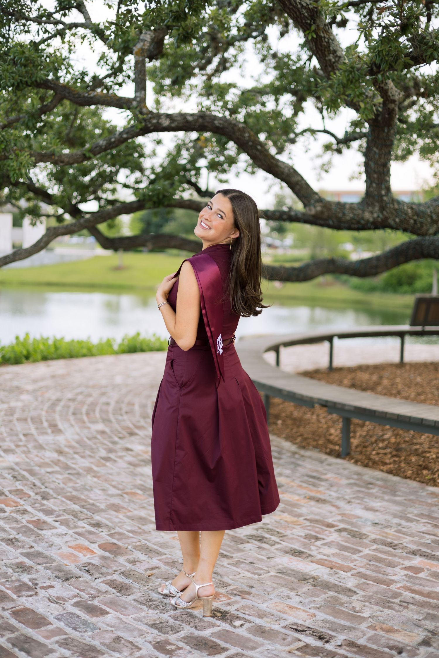 Smiling brunette woman in maroon Texas A&M graduation dress posing outdoors by a pond.