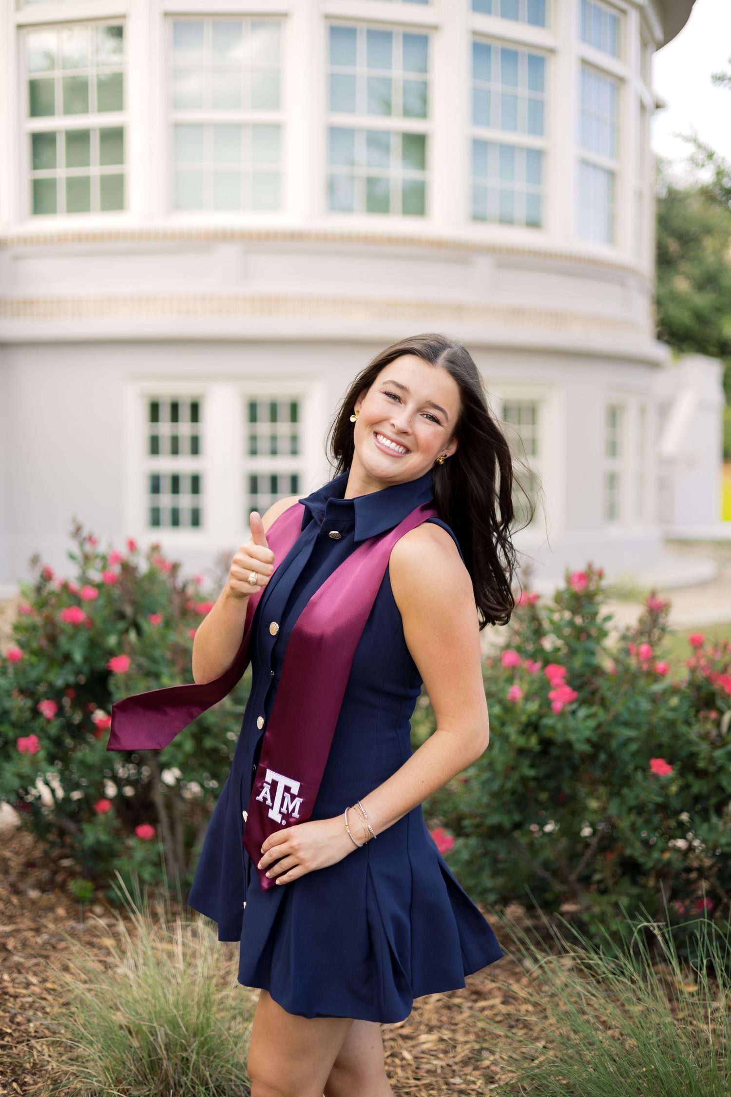 Happy woman in navy dress with Texas A&M maroon graduation stole giving thumbs up outdoors near rose bushes.