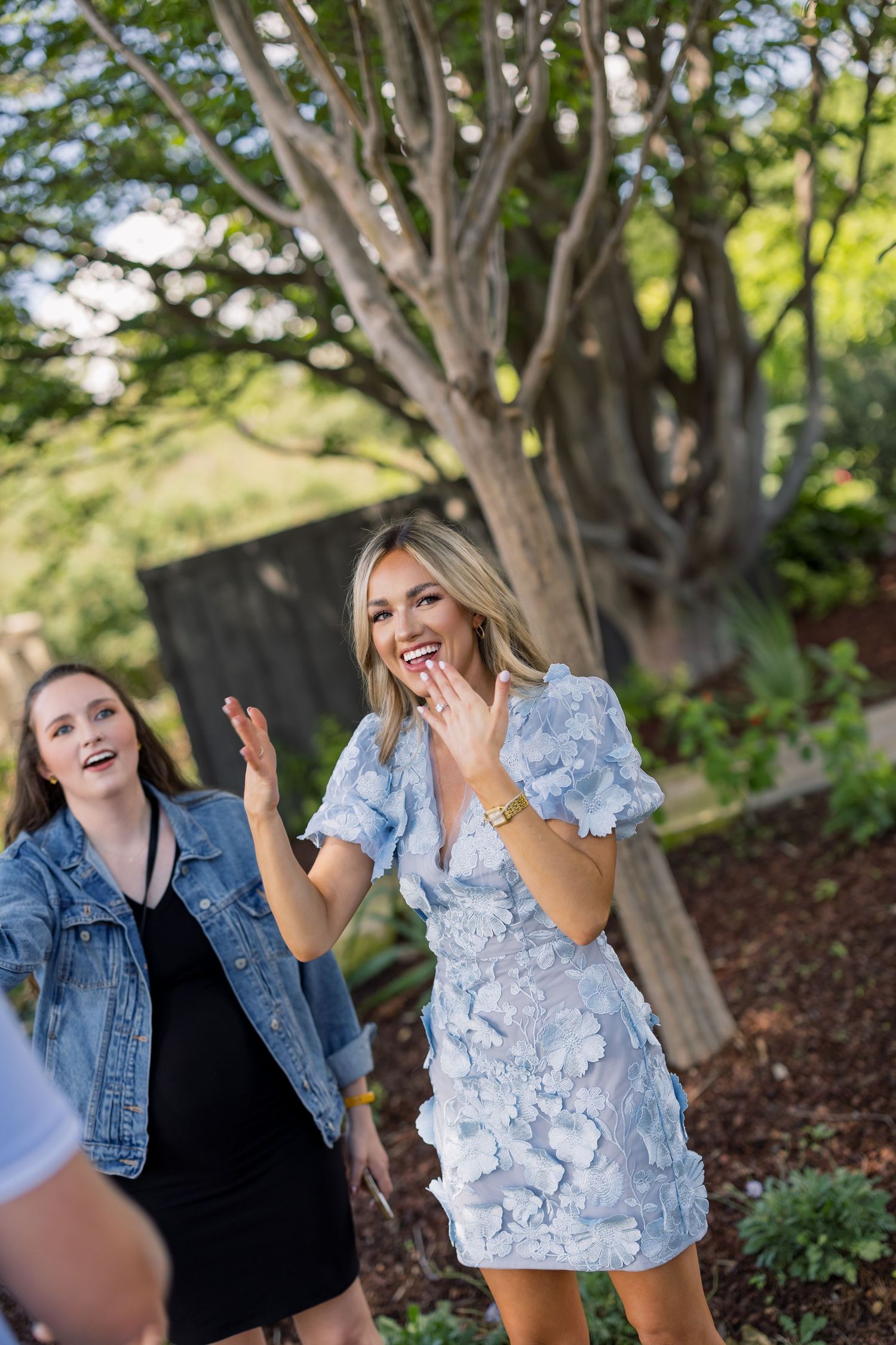 Two women laughing outdoors, one in a blue floral dress and one in a denim jacket, enjoying a garden party.