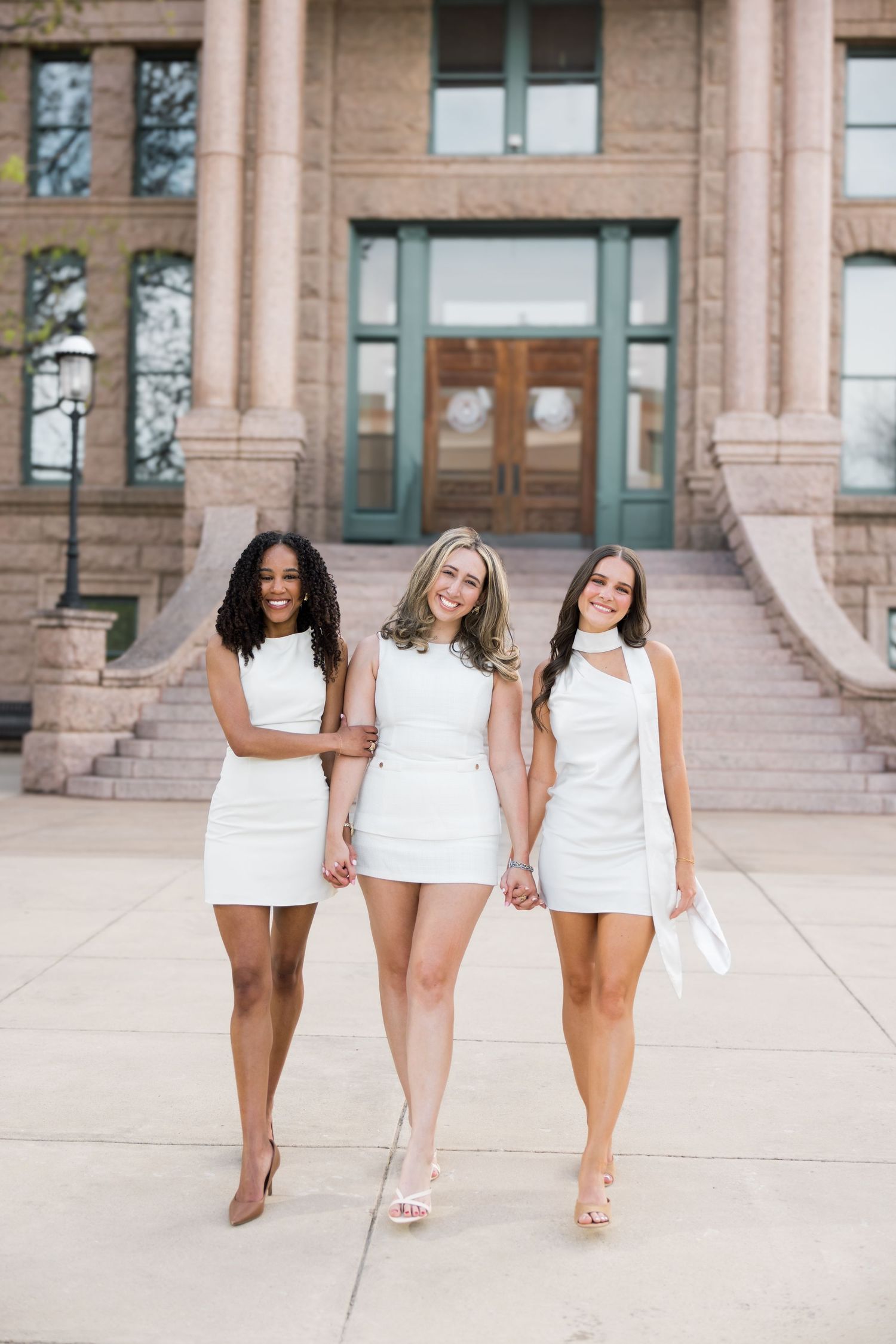 Three smiling women in white mini dresses walking together in front of a stone university building.