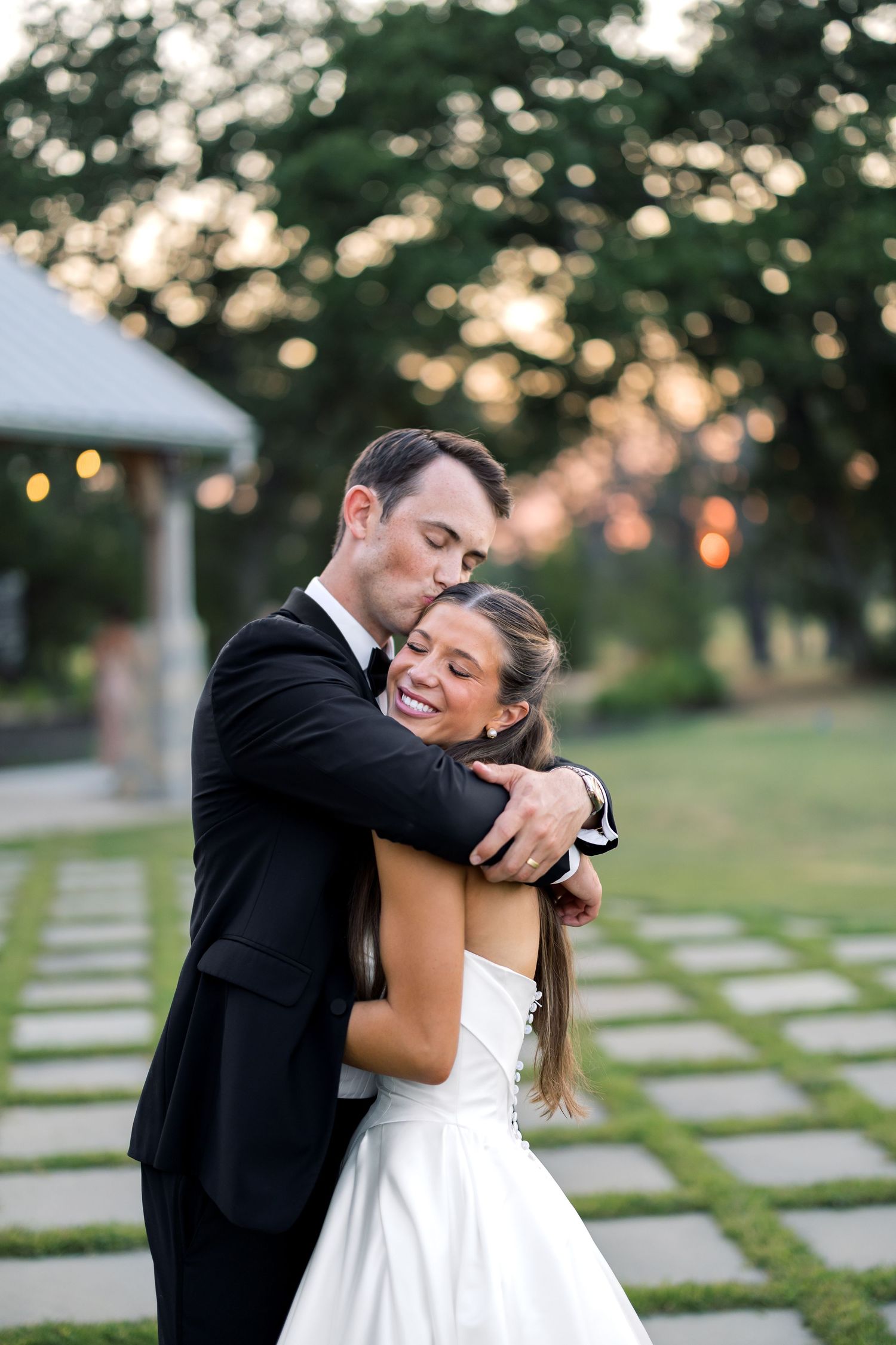 Groom in black suit embraces smiling bride in white gown outdoors at golden hour wedding reception.