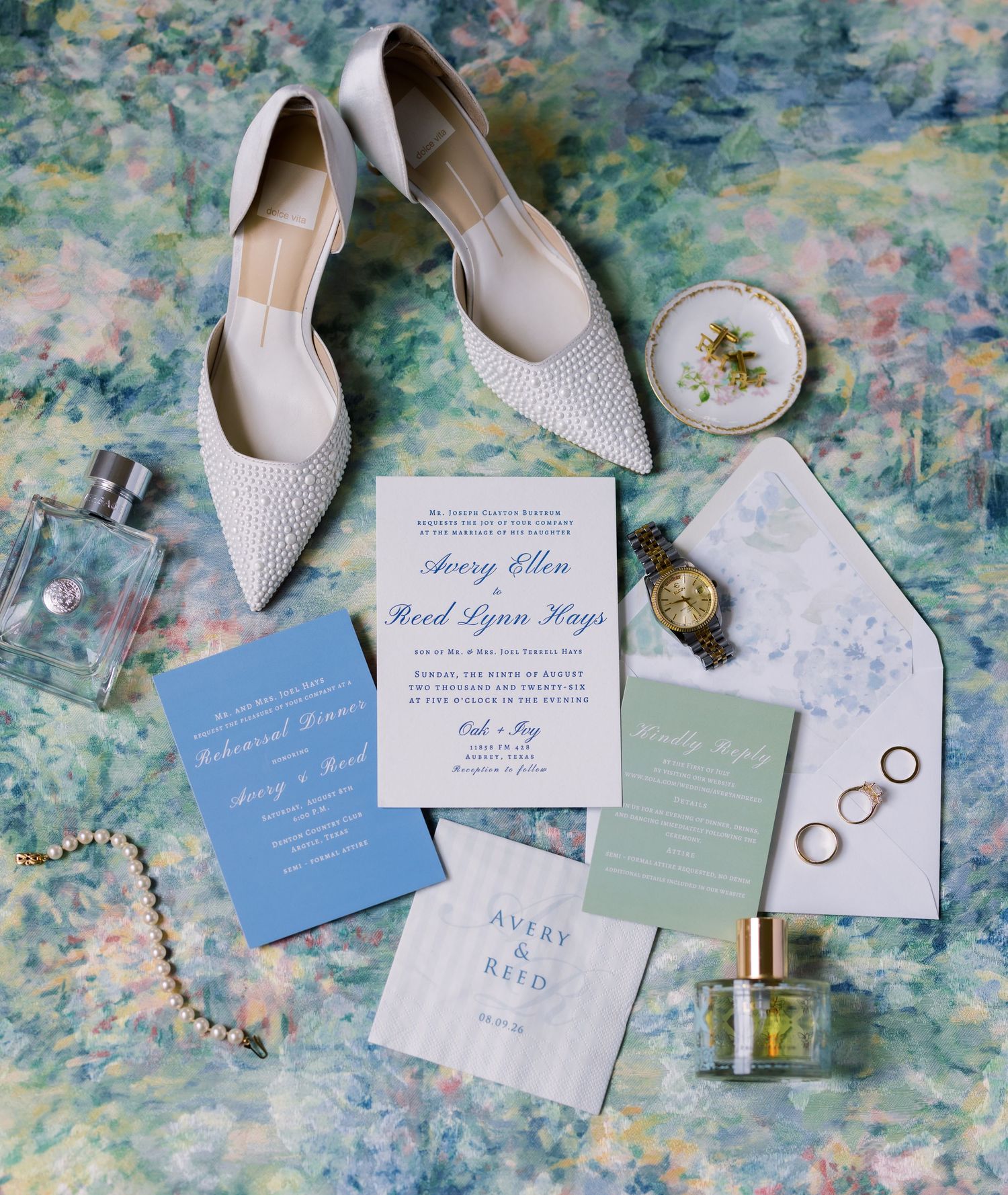 Elegant wedding flat lay with pearl heels, blue invitations, gold watch, rings, perfume, and floral stationery on watercolor backdrop.