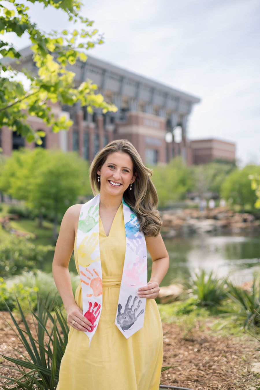 Smiling woman in yellow dress wearing colorful handprint scarf, posing outdoors near a pond and brick building.