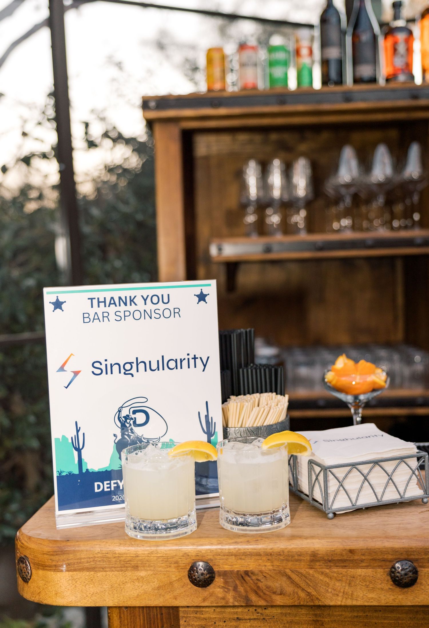 Singhularity bar sponsor sign beside two cocktails with lemon garnishes at an outdoor event bar.