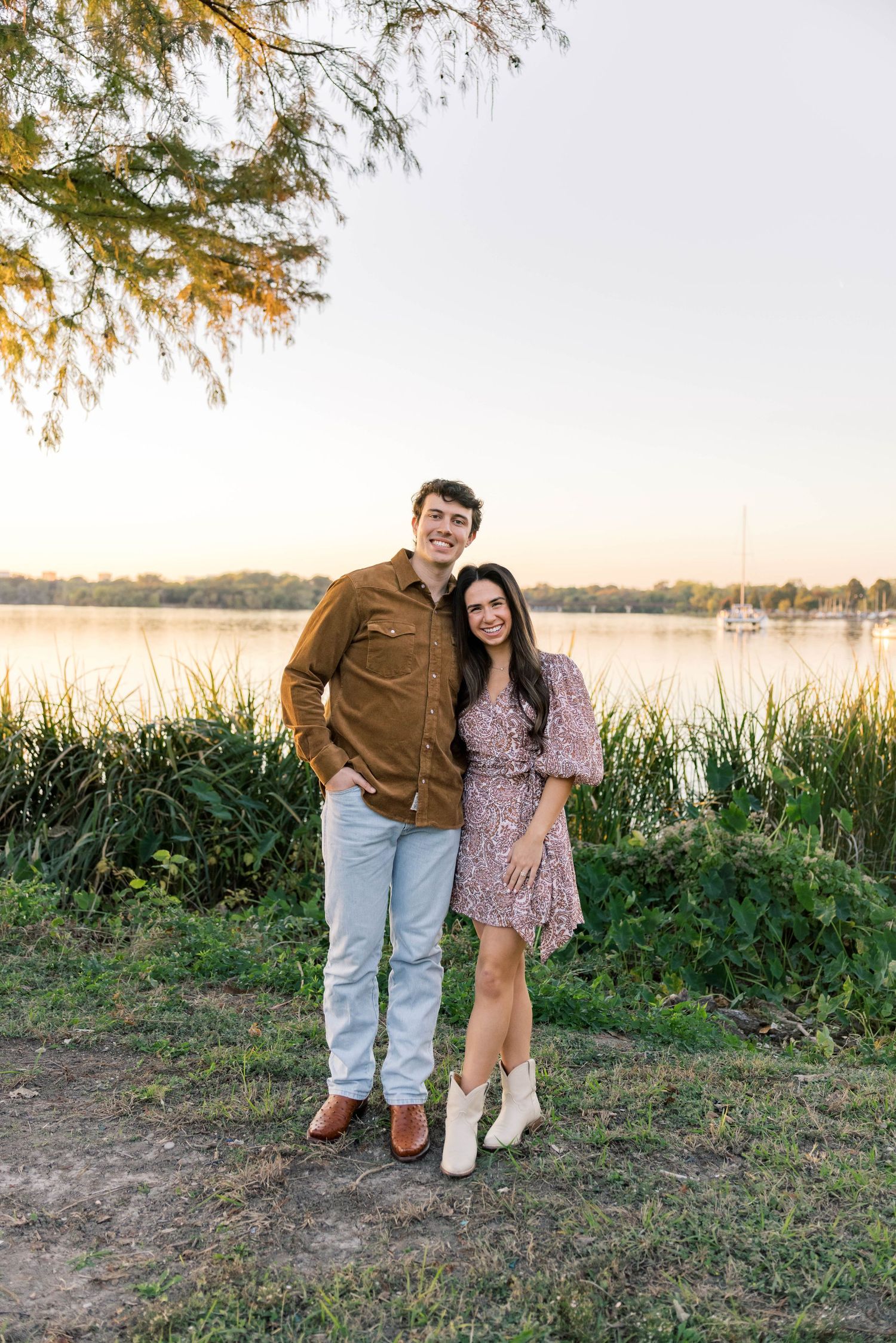Engaged couple posing together near a serene lake at sunset in a lush green park.