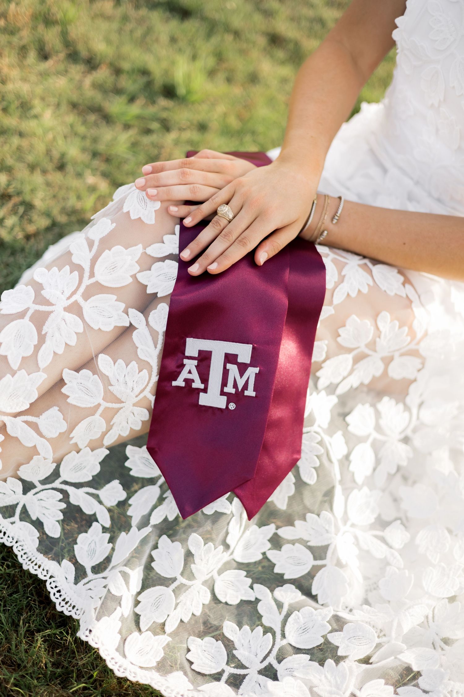 Close-up of hands resting on white lace dress with maroon Texas A&M graduation stole, wearing an engagement ring.