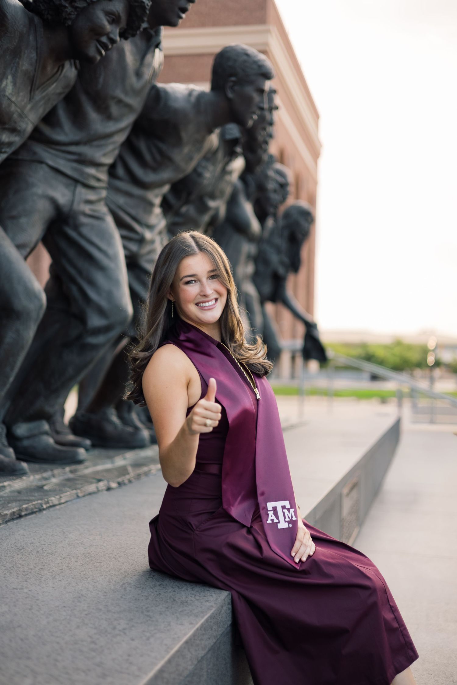 Smiling Texas A&M grad gives thumbs up in maroon graduation stole beside bronze statues.
