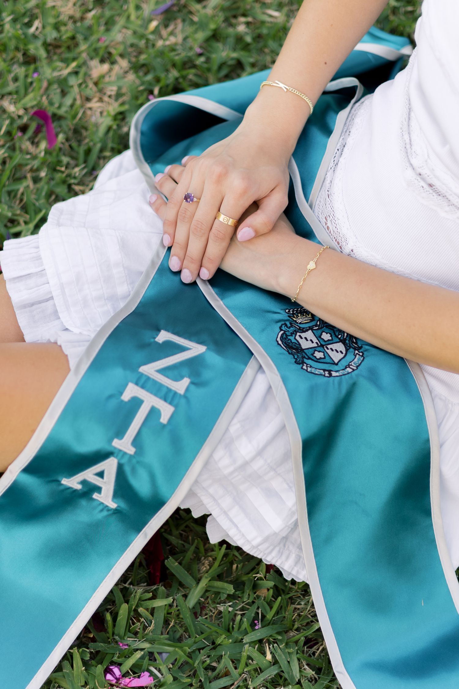 Close-up of hands resting on a teal ZTA sorority graduation sash laid on green grass.