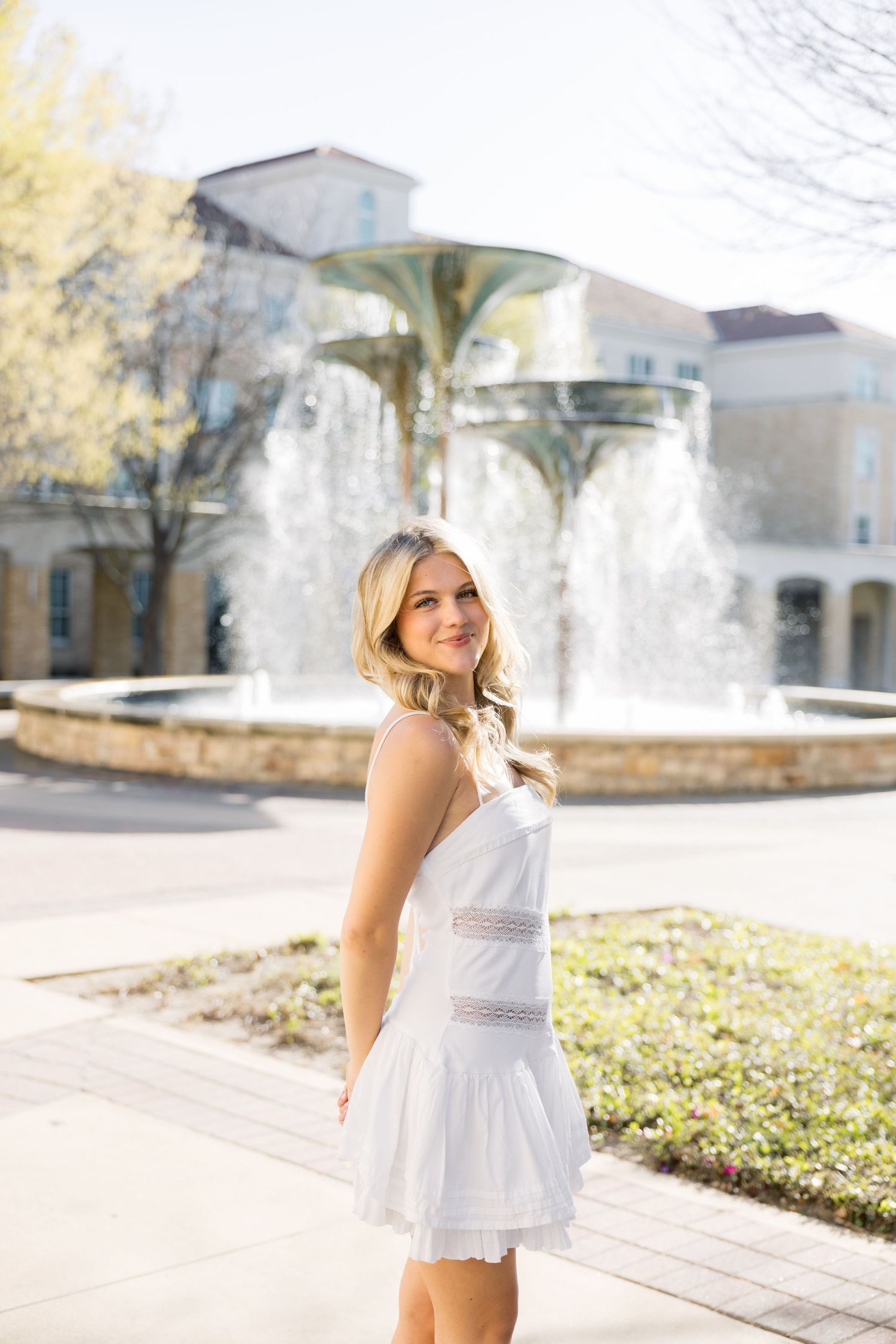 Blonde woman in white lace dress smiles over shoulder near ornate fountain on college campus.