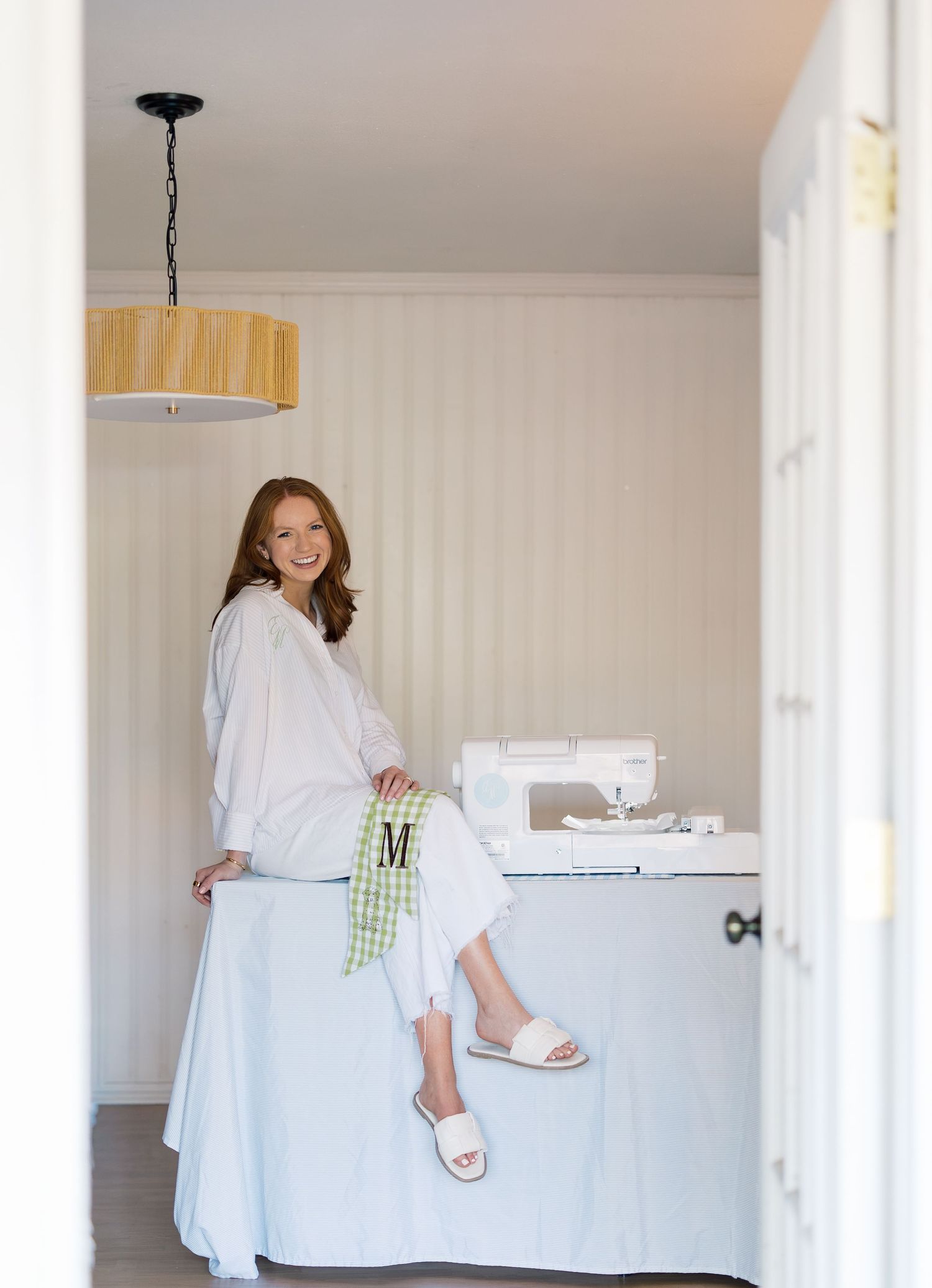 Woman in white outfit sits on a sewing table beside a sewing machine in a bright, cozy craft room with a rattan pendant light.