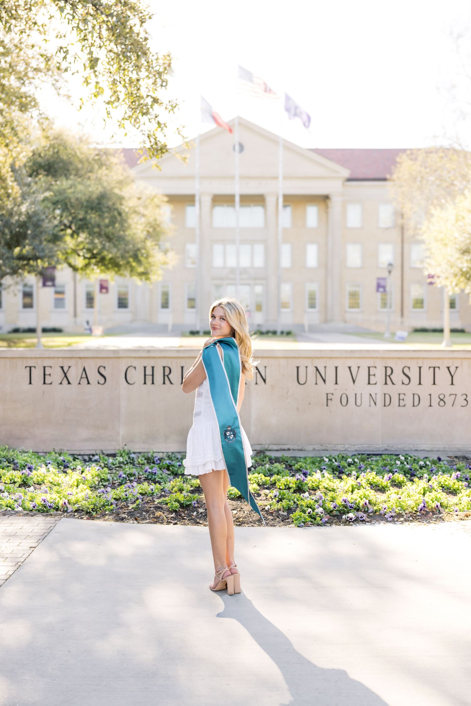 Blonde graduate in white dress with teal sash posing at Texas Christian University Founded 1873 sign.