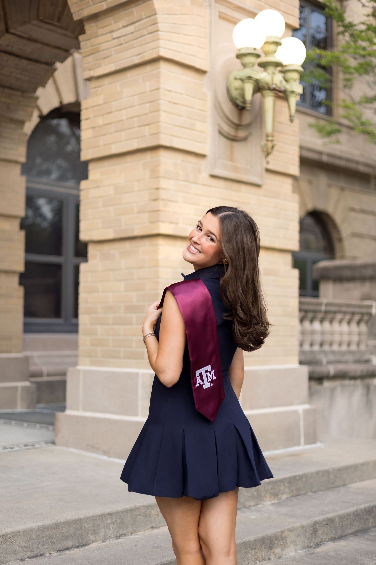 Young woman in navy dress with Texas A&M maroon graduation stole posing outside a stone building.