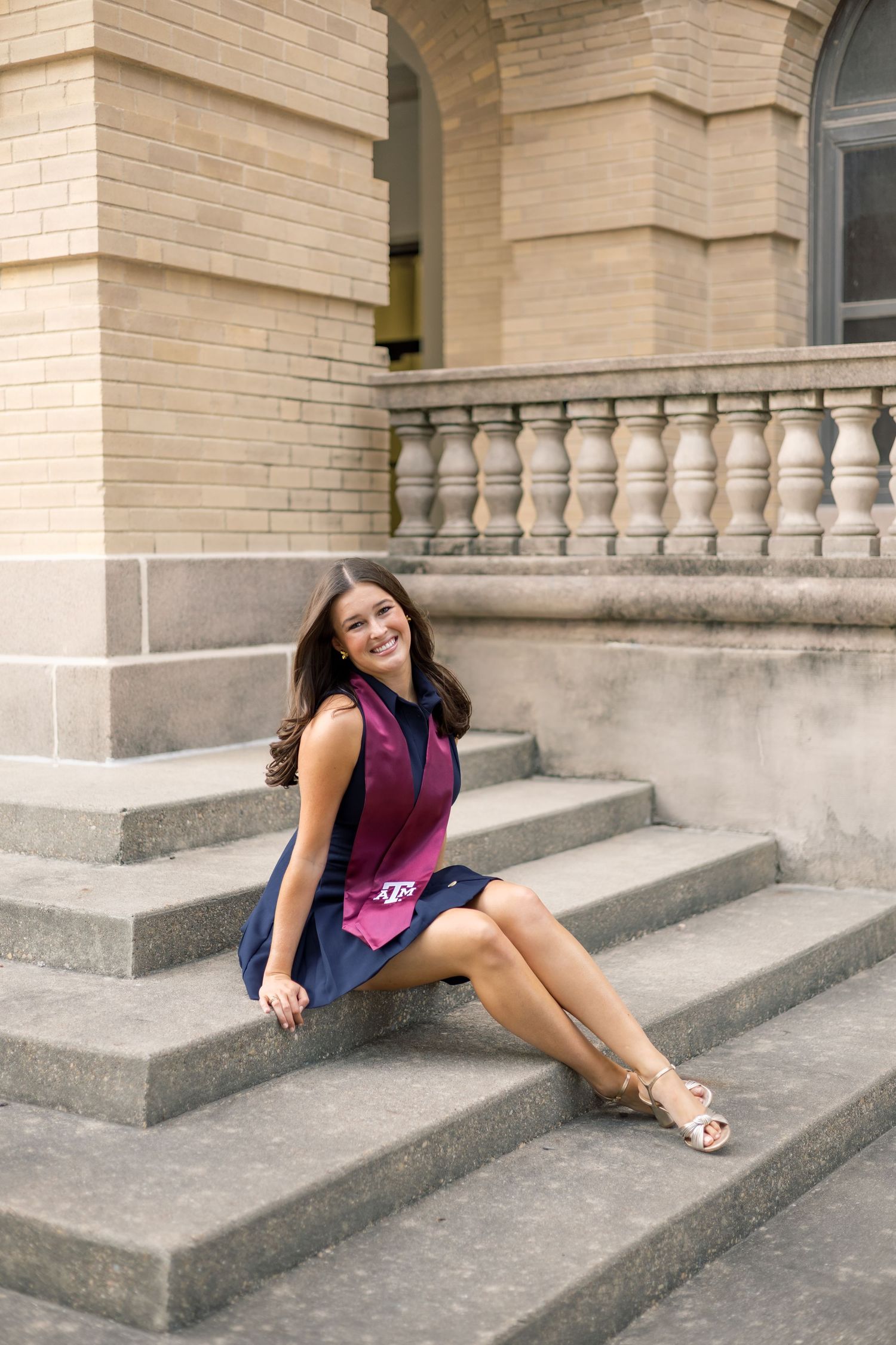 Smiling young woman in navy and maroon Texas A&M dress sitting on stone steps of a classic building.