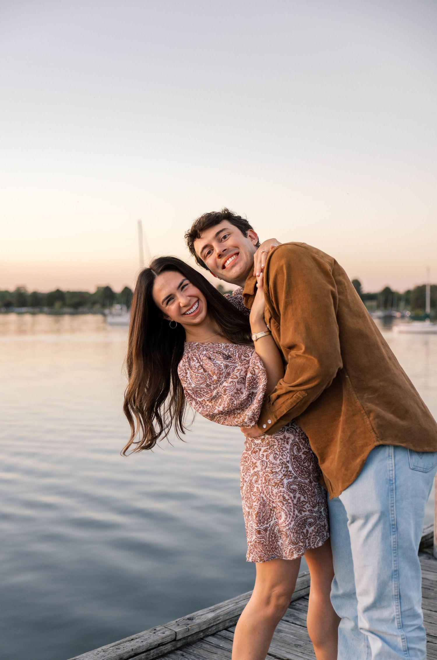 A smiling couple embraces on a wooden dock at sunset with calm water and sailboats in the background.