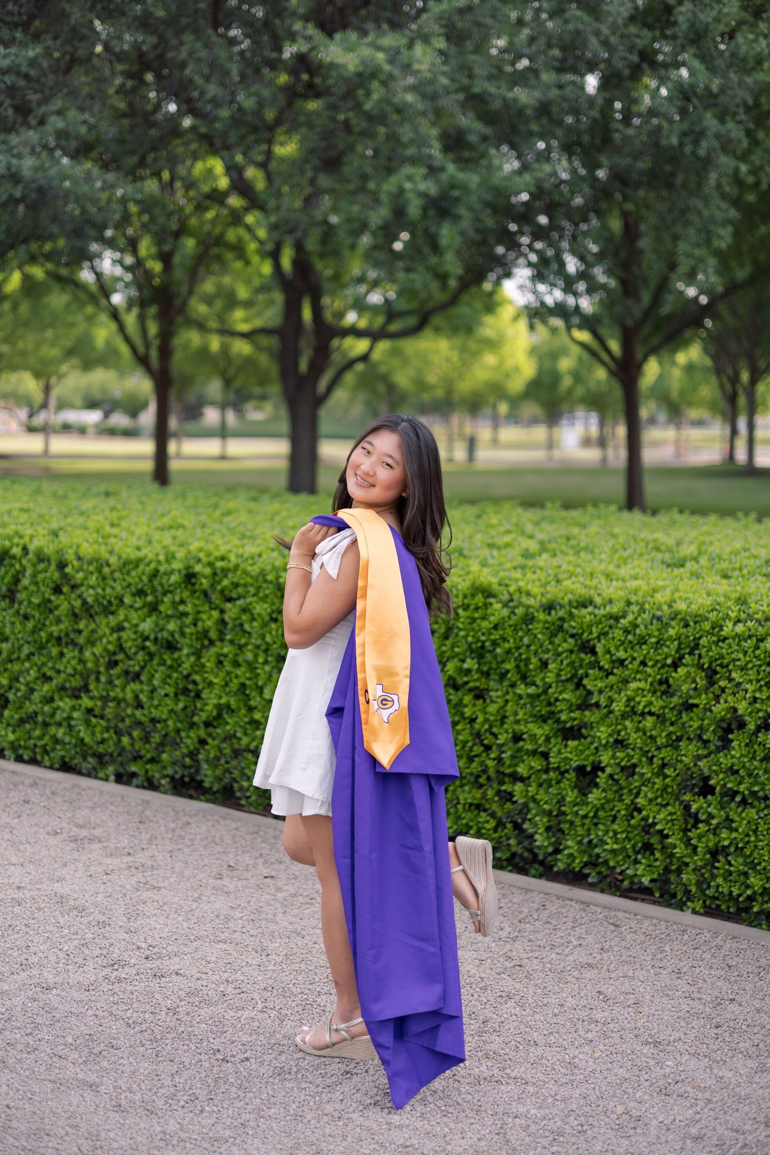 Young woman in white dress holding purple graduation gown and gold stole, posing outdoors near green hedges.