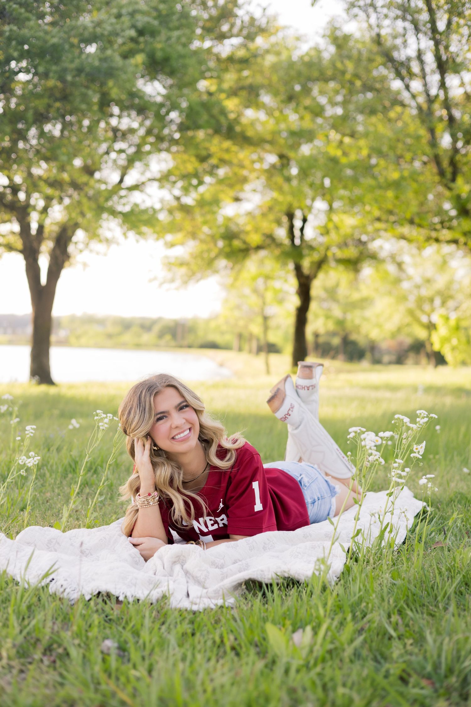 Smiling blonde woman lying on a blanket in a green park wearing a maroon jersey and jeans on a sunny day.