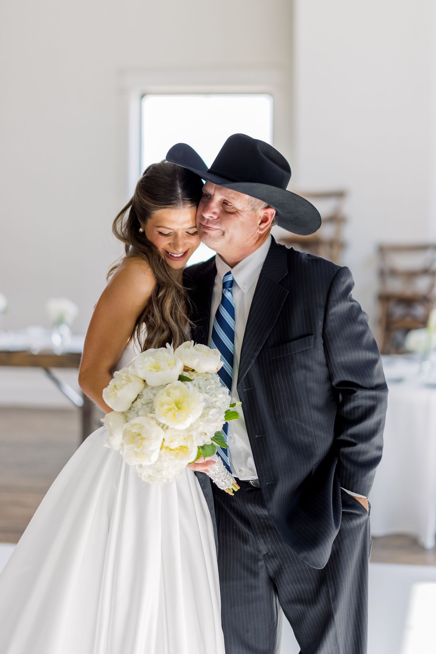 Bride holding white peony bouquet smiles as older man in black cowboy hat and suit kisses her cheek at wedding reception.