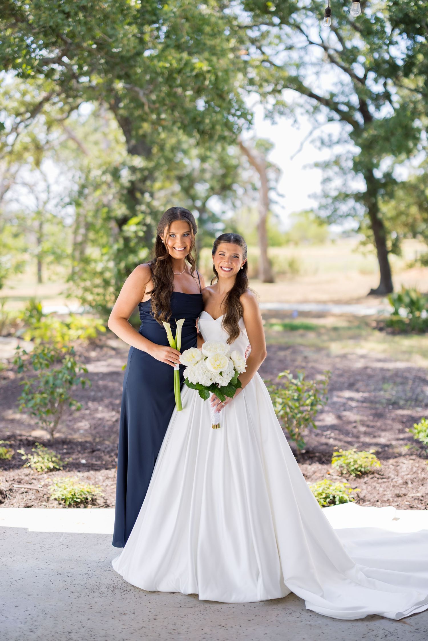 Bride in white gown and bridesmaid in navy dress holding white calla lily bouquets outdoors.