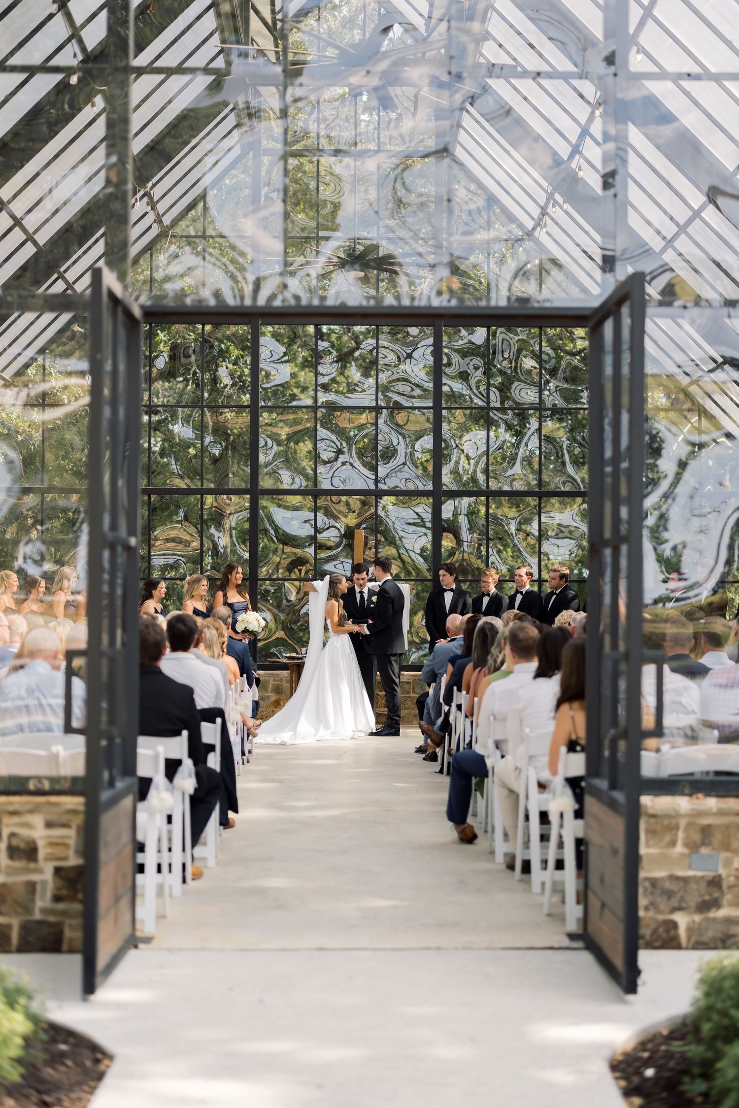 Elegant greenhouse wedding ceremony with bride and groom at altar, guests seated in white chairs, lush botanical backdrop.