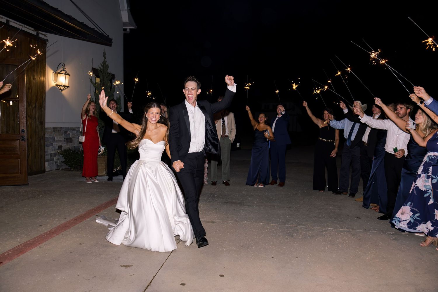 Bride and groom exit wedding reception through sparkler tunnel surrounded by guests at night.