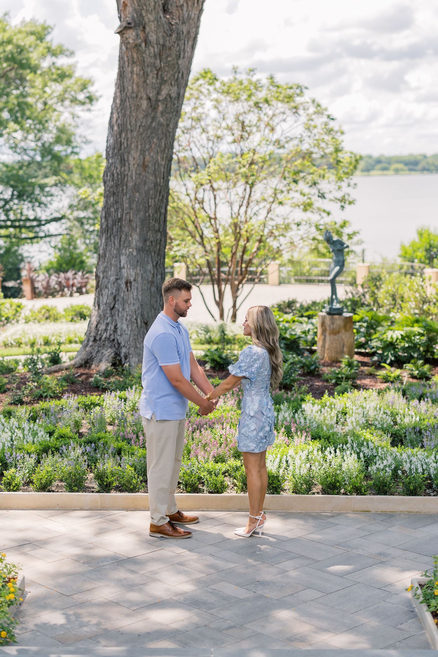 Couple holding hands in a lush garden with a large tree, statue, and waterfront view on a sunny day.