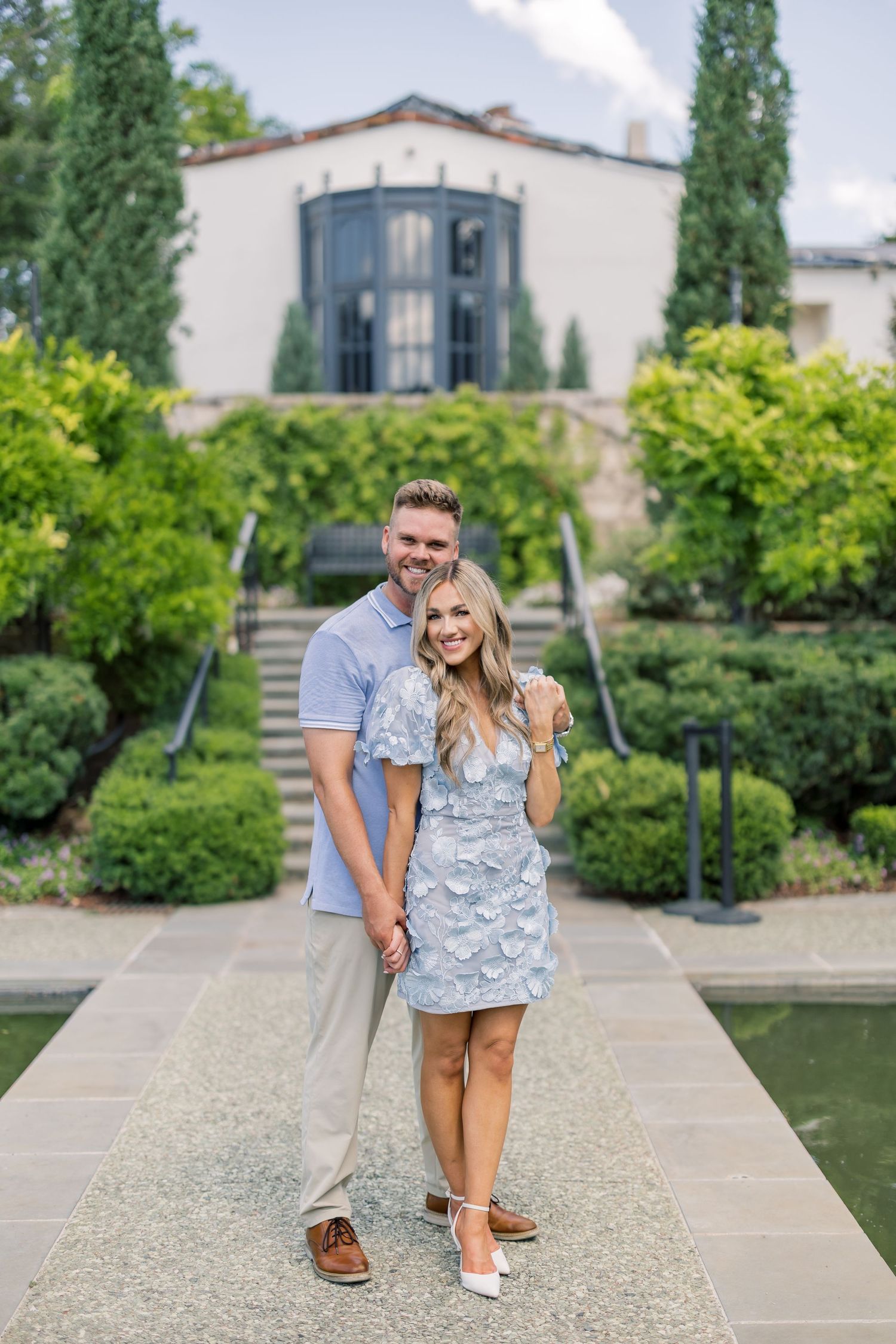 Smiling couple posing in formal garden with lush greenery and elegant white building backdrop on sunny day.