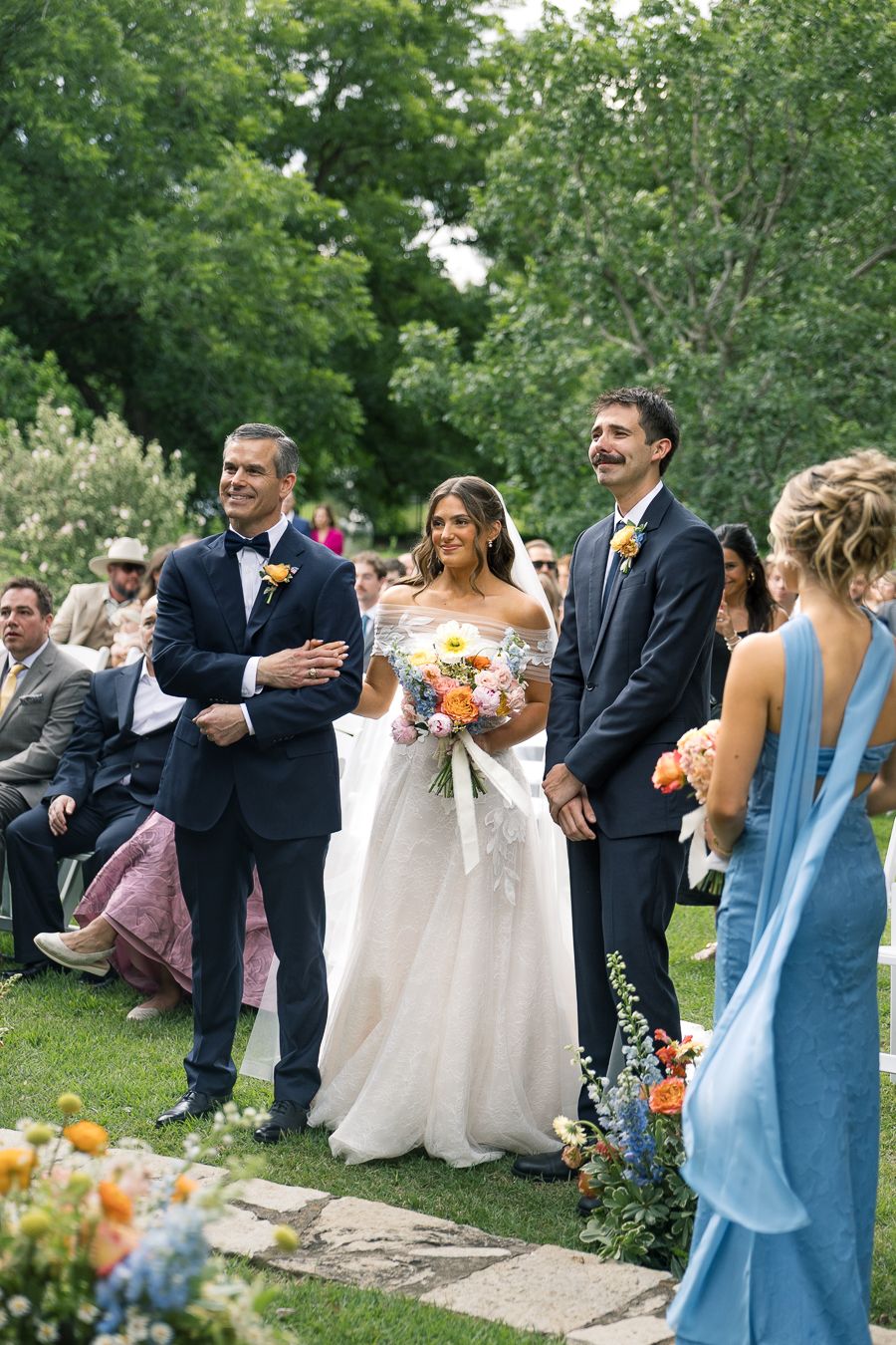 Bride in white gown holds colorful bouquet, flanked by two men in suits, during outdoor wedding ceremony.