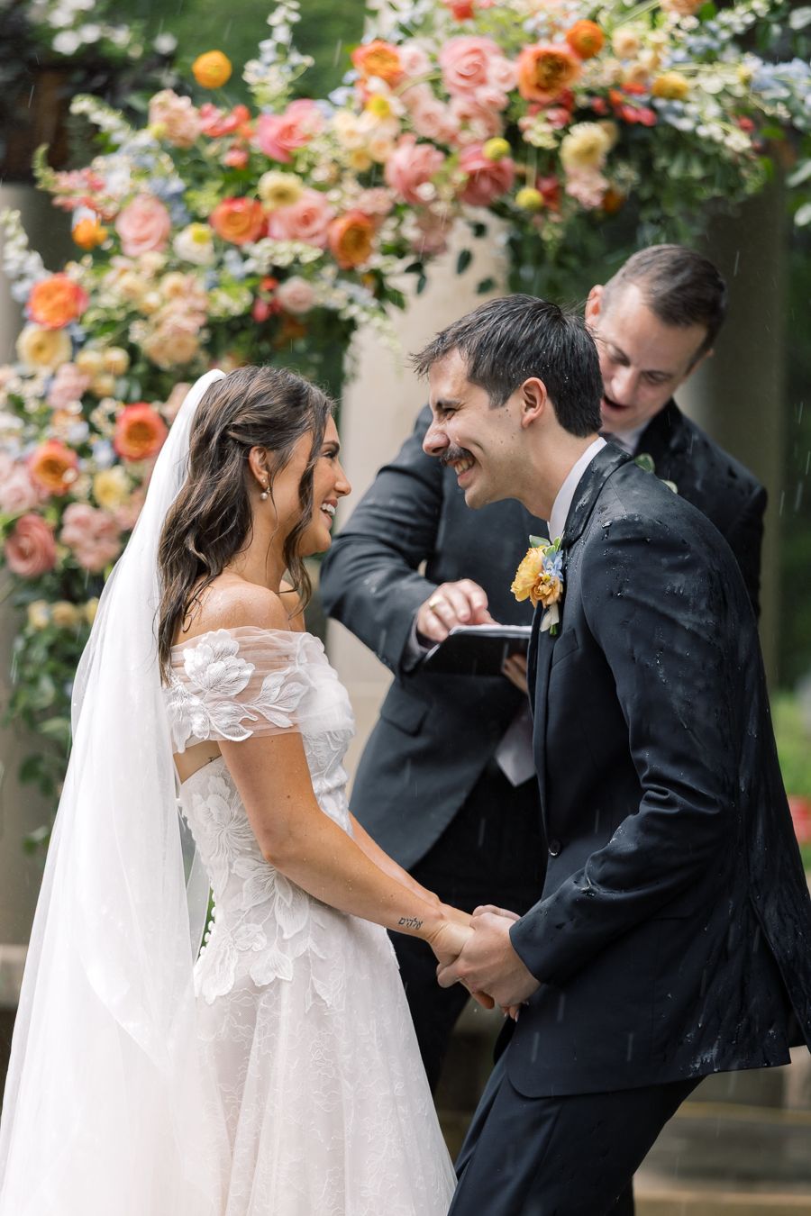 Bride and groom laughing joyfully during outdoor wedding ceremony beneath colorful floral arch with officiant.