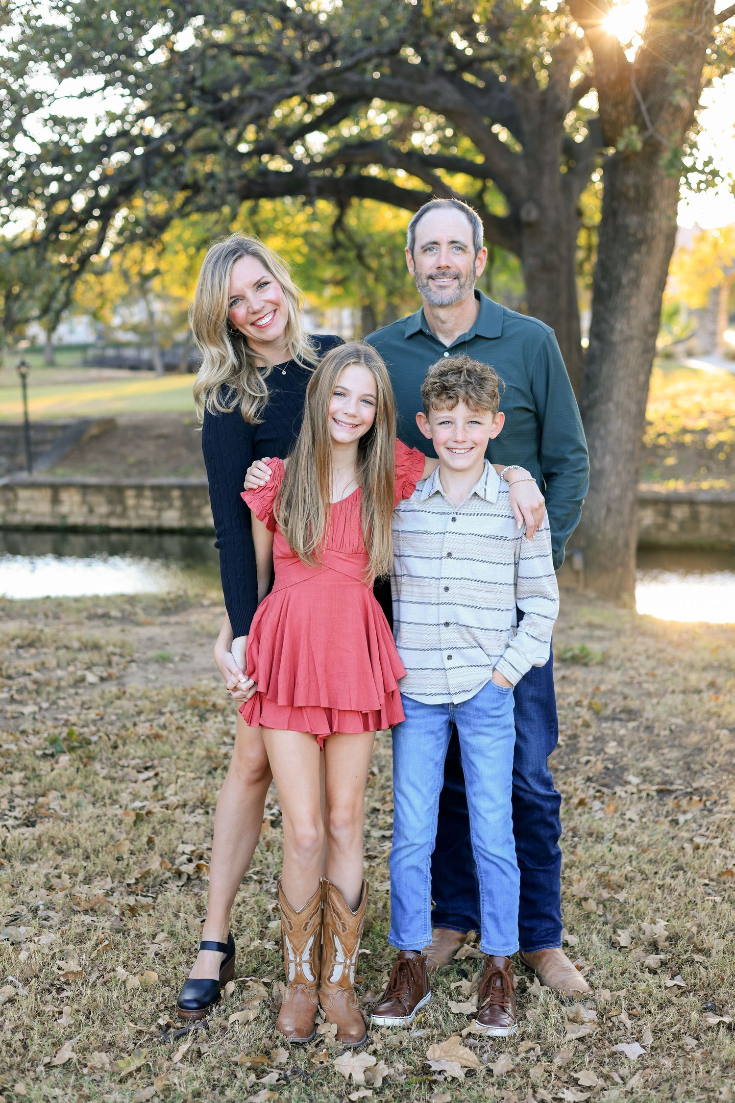 Family of four posing outdoors near a pond and oak tree during golden hour in a park setting.