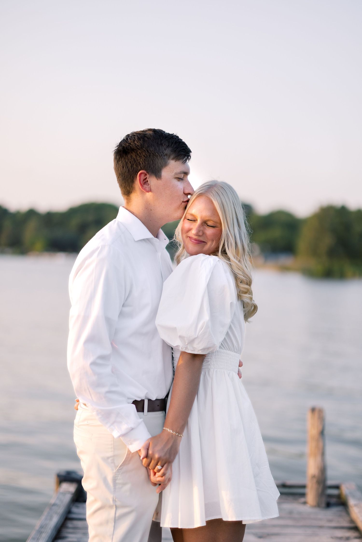 Couple in white outfits sharing a kiss on a lakeside dock at golden hour.