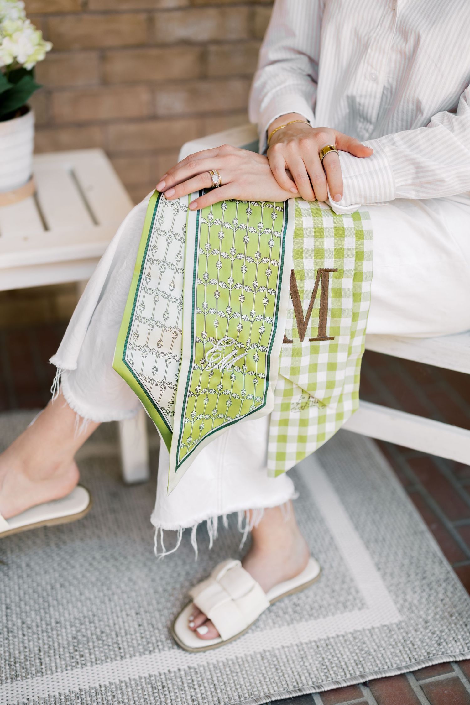 Woman sitting outdoors holding a green gingham monogrammed M silk scarf on her lap, wearing white sandals.
