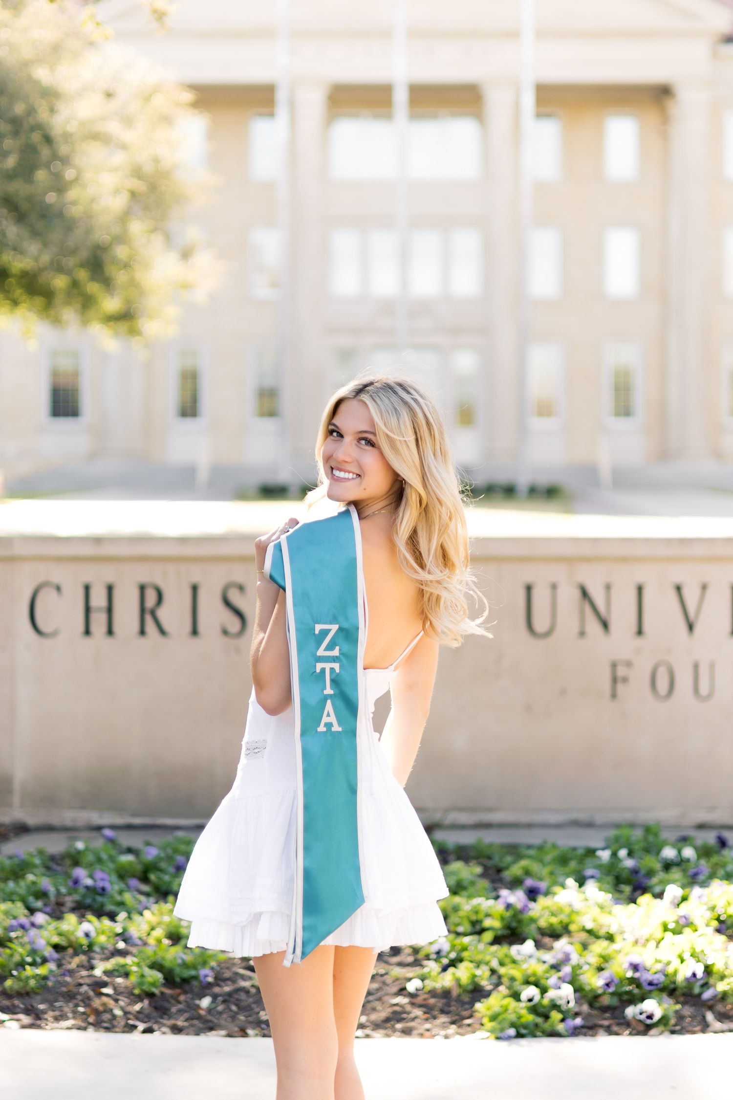 Smiling blonde woman in white dress holding teal ZTA graduation sash in front of Christian University building.