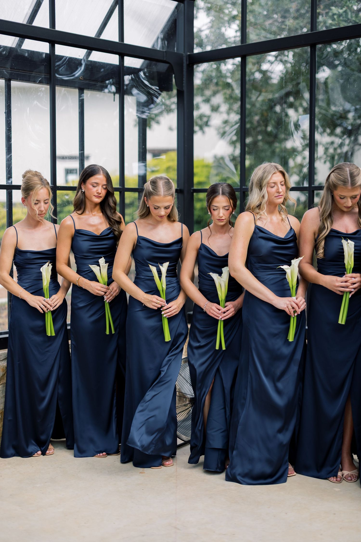 Six bridesmaids in navy satin gowns holding white calla lilies, standing in an elegant glass-walled venue.