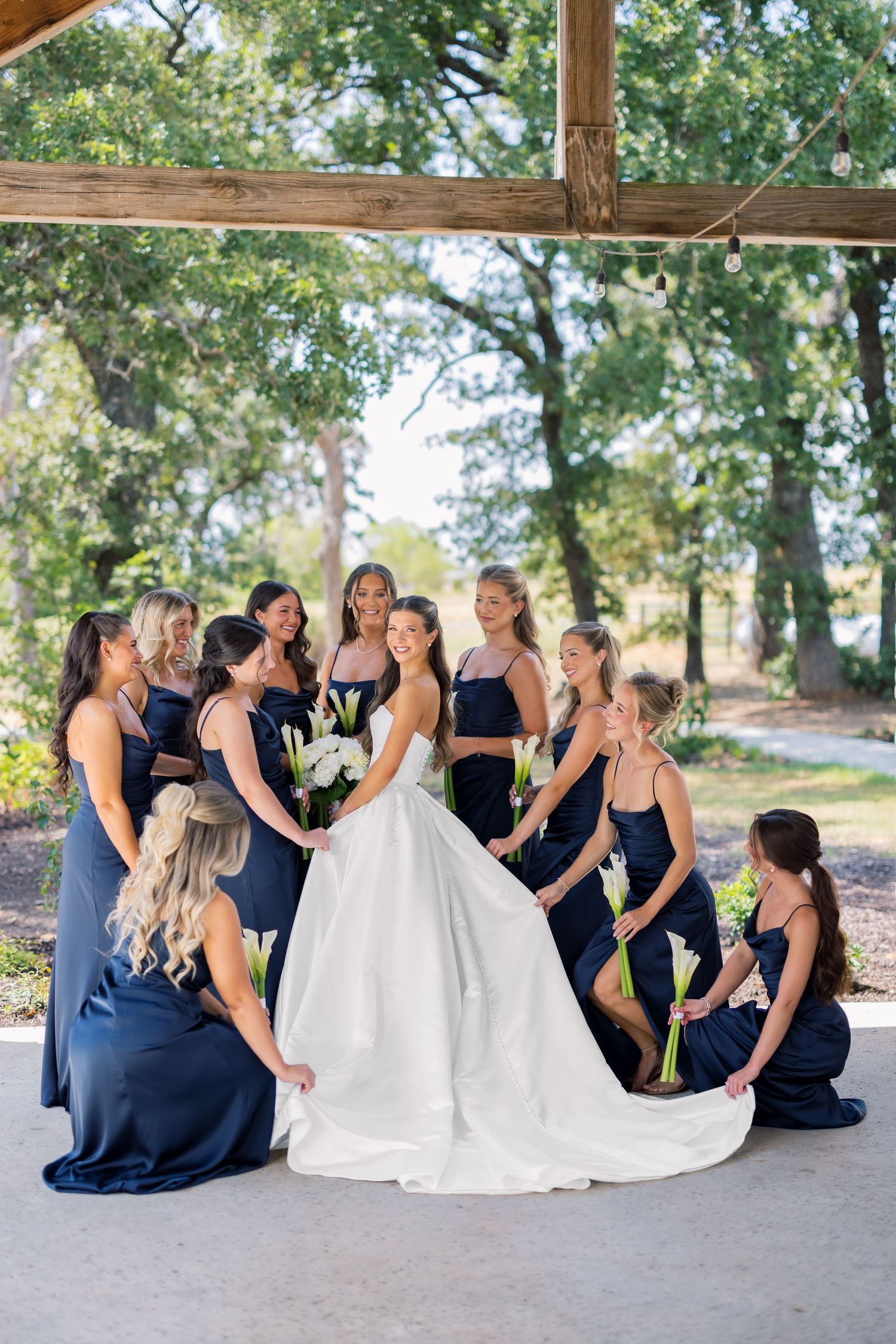 Bride in white gown surrounded by bridesmaids in navy blue dresses holding white calla lilies under a rustic outdoor pavilion.