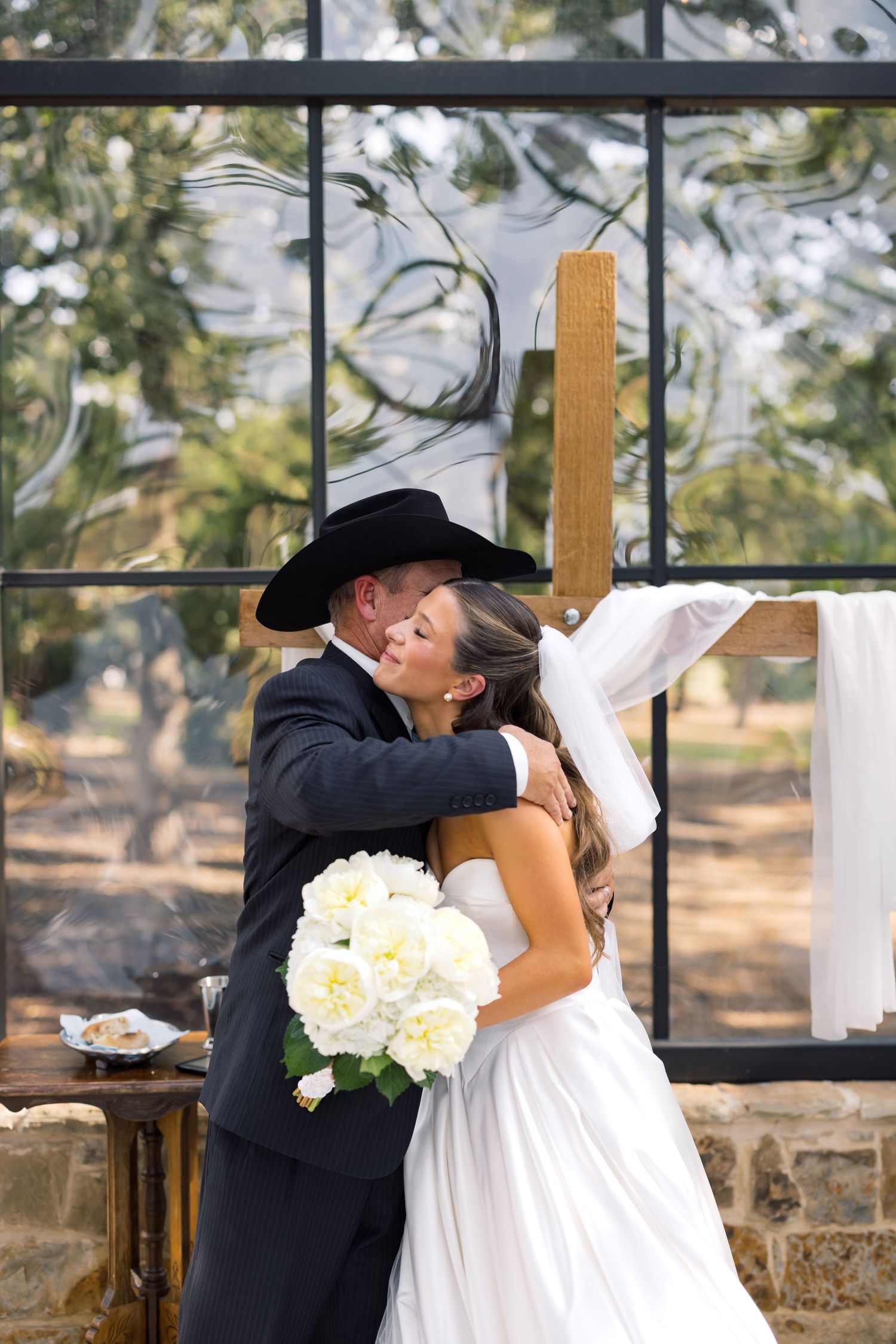 Bride and groom share first kiss at rustic outdoor wedding ceremony, bride holding white rose bouquet, groom wearing black cowboy hat.