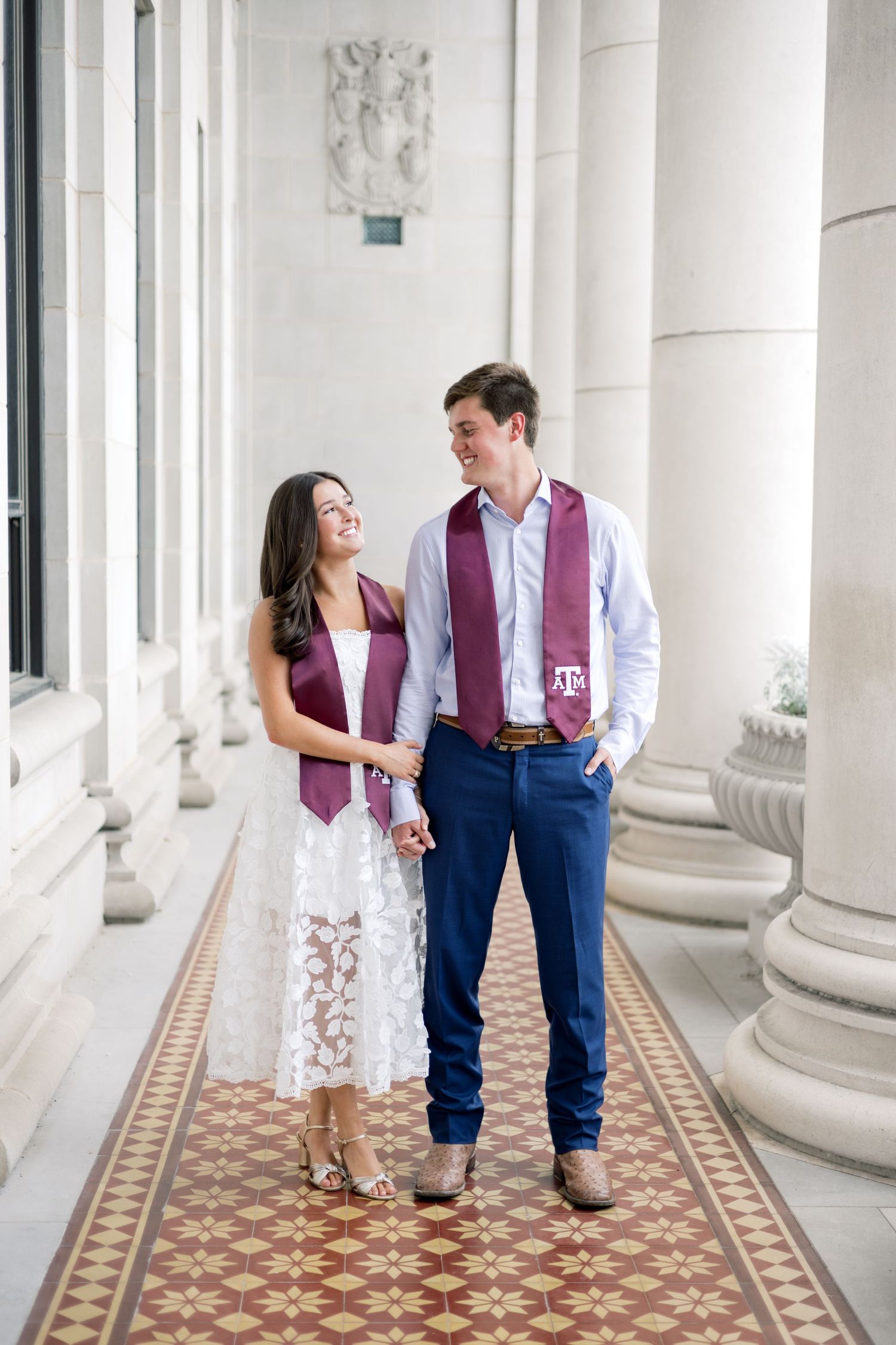 Couple in Texas A&M maroon graduation stoles holding hands and smiling in a grand columned hallway with decorative tile.