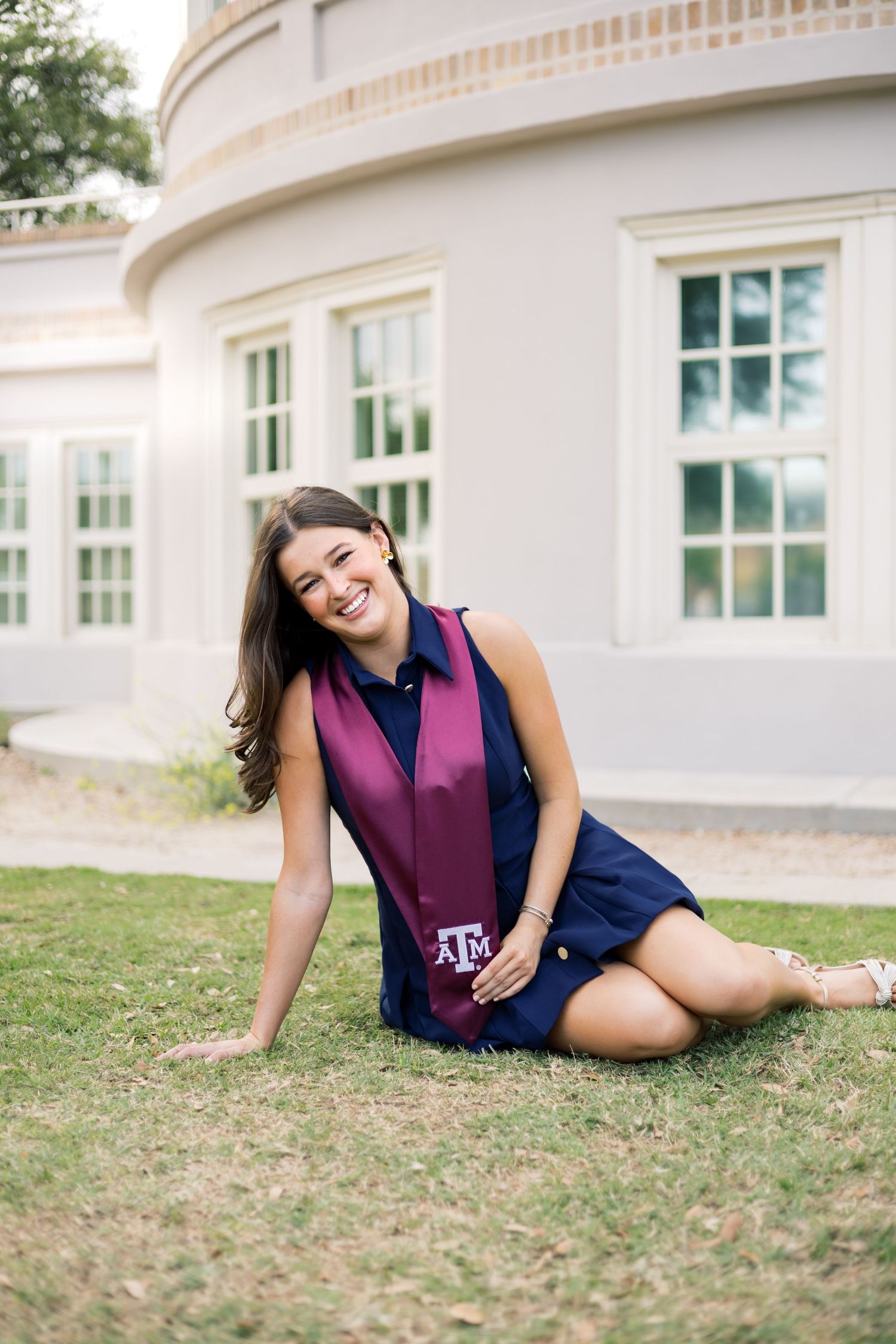 Smiling Texas A&M graduate poses on grass in navy dress with maroon graduation stole near campus building.