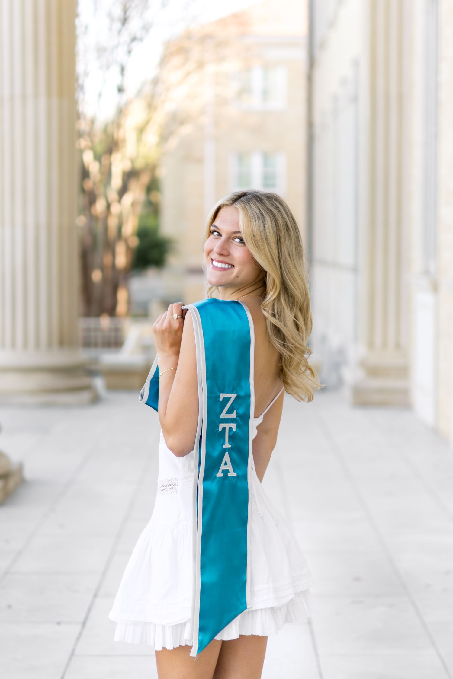 Smiling blonde woman in white dress holding a teal ZTA graduation sash between campus columns.