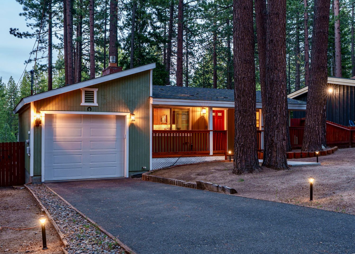 String lights illuminate a cozy mountain cabin driveway surrounded by tall pine trees at dusk.