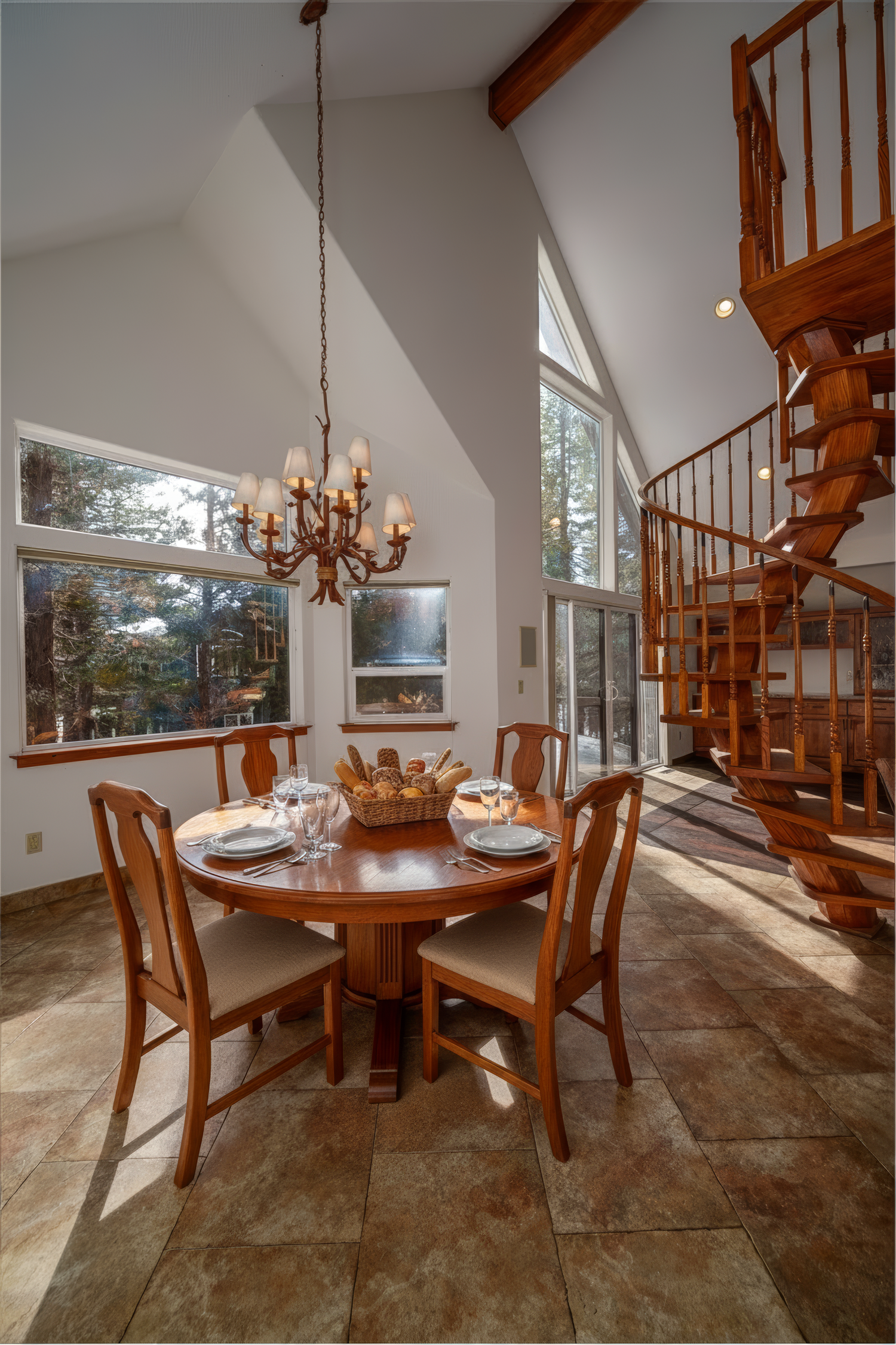 Elegant dining room with spiral staircase, chandelier, and large windows overlooking forest in a vaulted ceiling space.