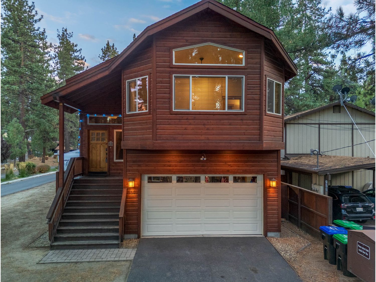 Modern two-story mountain cabin with dark wood siding, white garage door, and exterior lighting in a wooded setting.