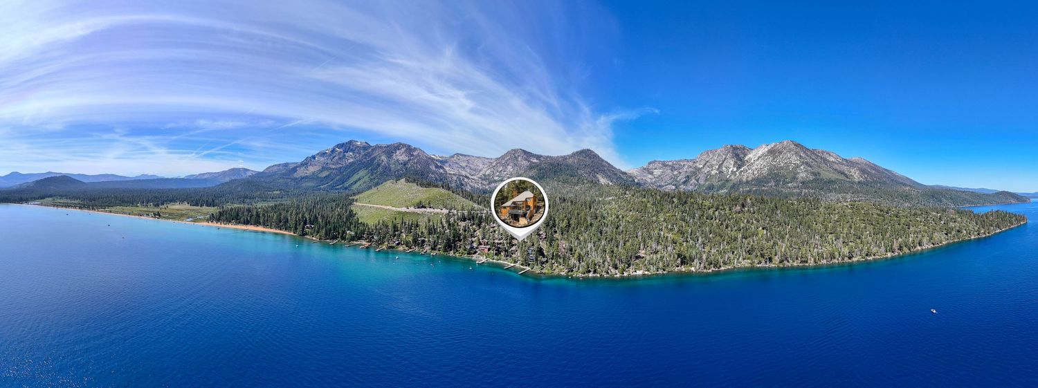 Aerial panoramic view of Lake Tahoe shoreline with crystal blue water, dense pine forests, and snow-capped Sierra Nevada mountains.