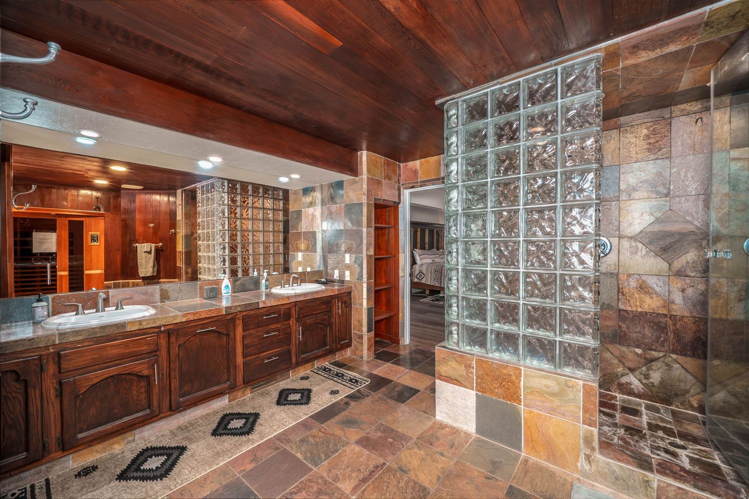 Rustic bathroom with wood ceiling, slate tile floors, dual vanity, and glass block shower wall.